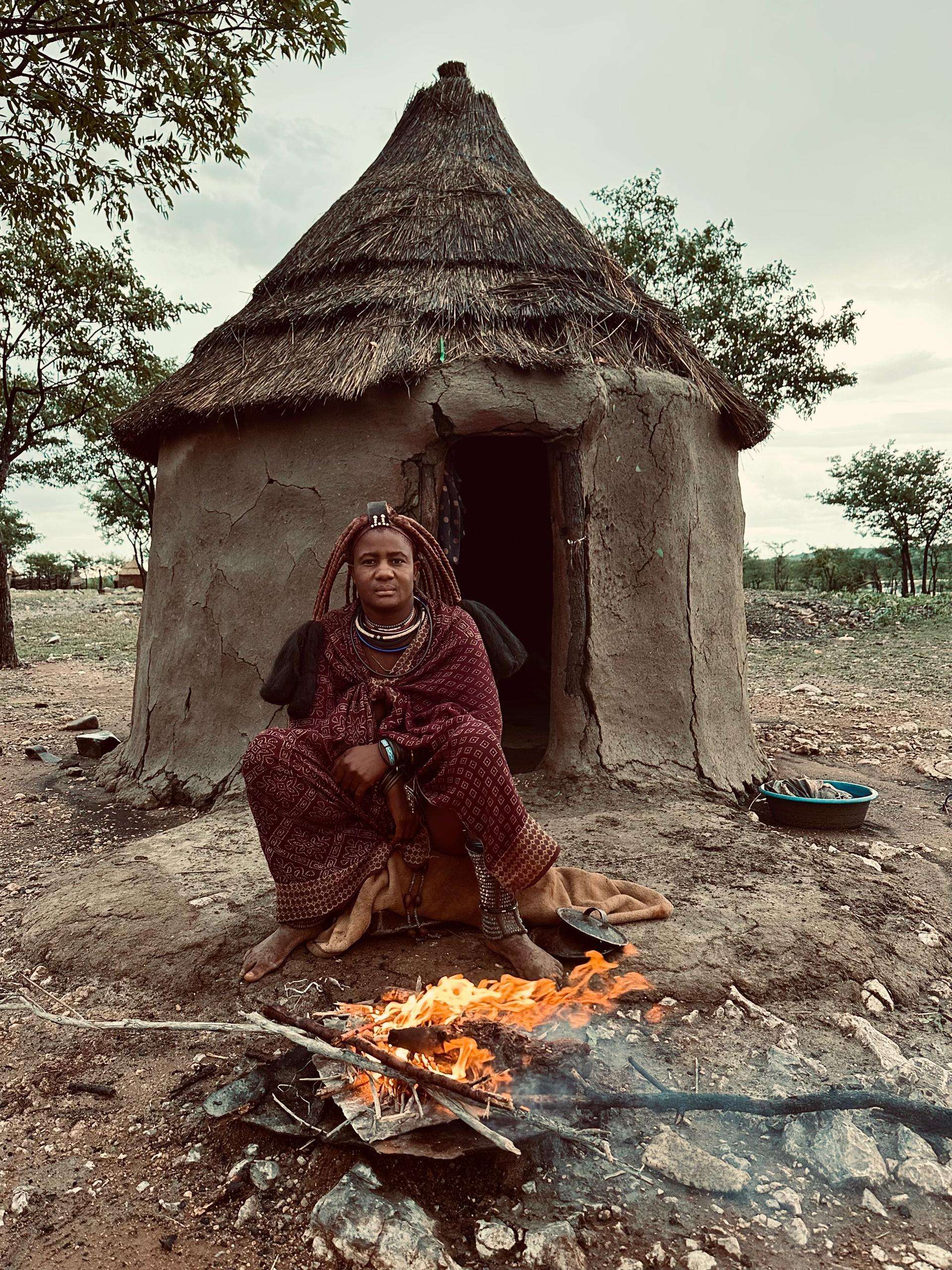 A Himba woman sitting outside a mud-and-thatch hut beside a lit fire