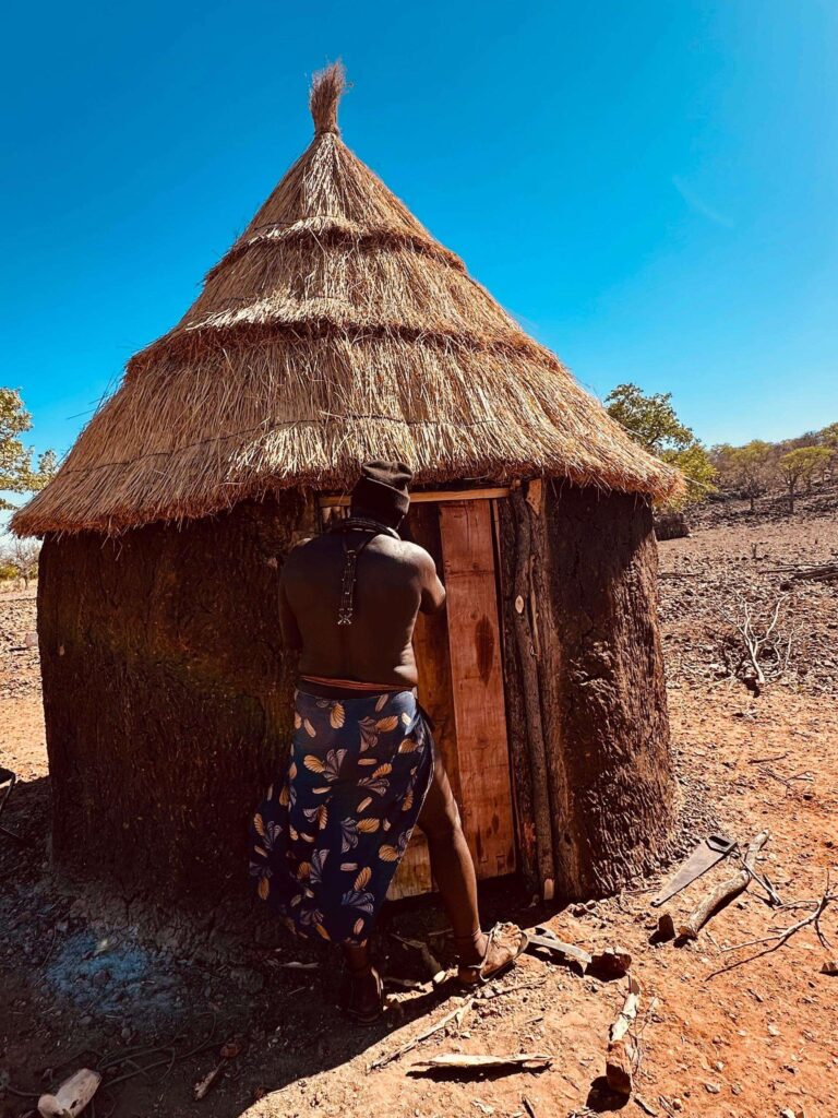 A Himba man opening the timber door of a thatched hut under a deep blue sky