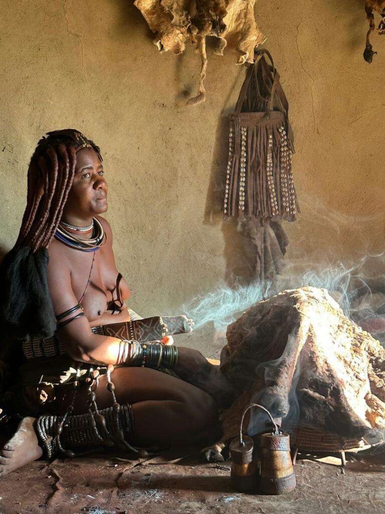 A Himba woman inside her hut beside a smoking fire, with skins and a beaded bag on the wall behind her