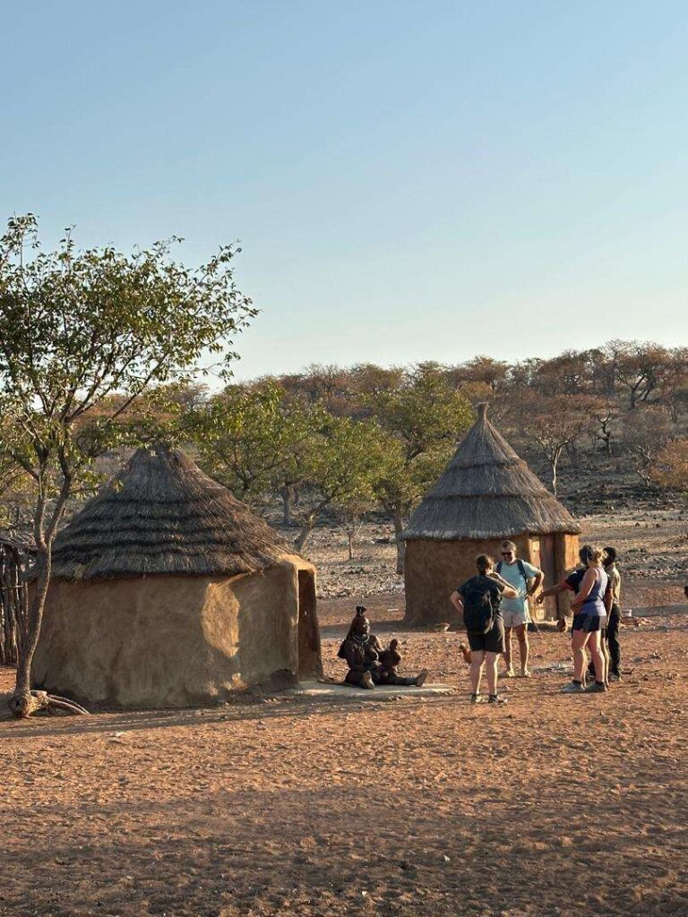 A small group of Two Farm guests standing between two thatched Himba huts in late afternoon light